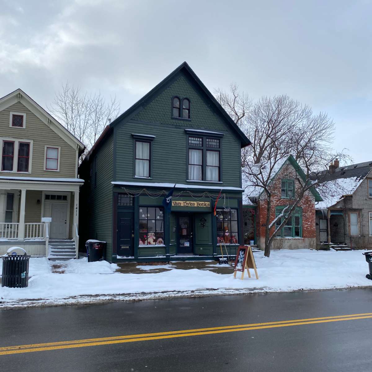 A two-story green house with a shop front and snow covering the ground stands alongside a snowy street with several other buildings in the background.