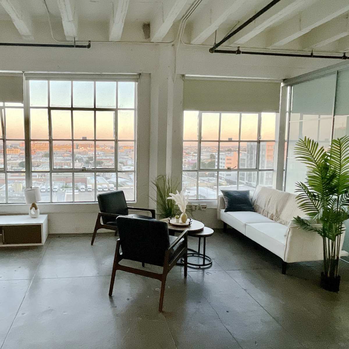 A modern, minimalist living space features large windows with an urban view at sunset, furnished with a sofa, chairs, and decorative plants.