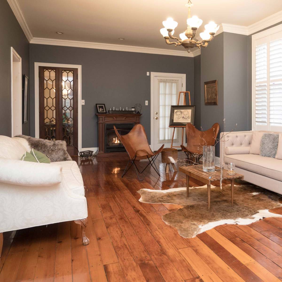A living room with wooden floors, a beige couch, a glass coffee table, and a fireplace, illuminated by a chandelier.