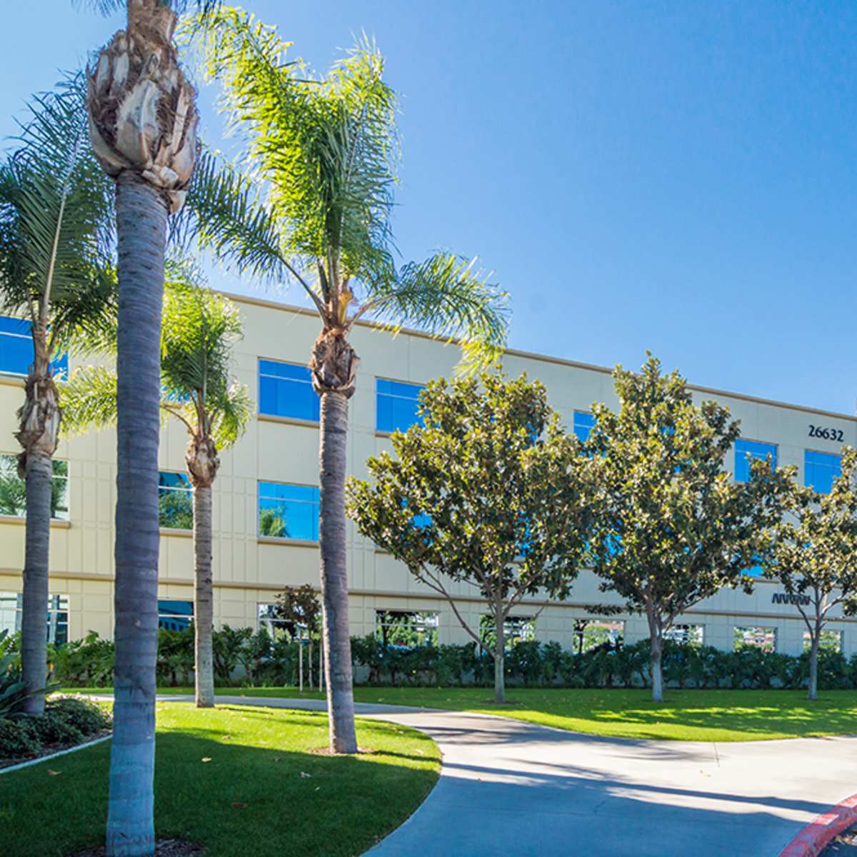 A modern office building with large glass windows, surrounded by palm trees and landscaped greenery.