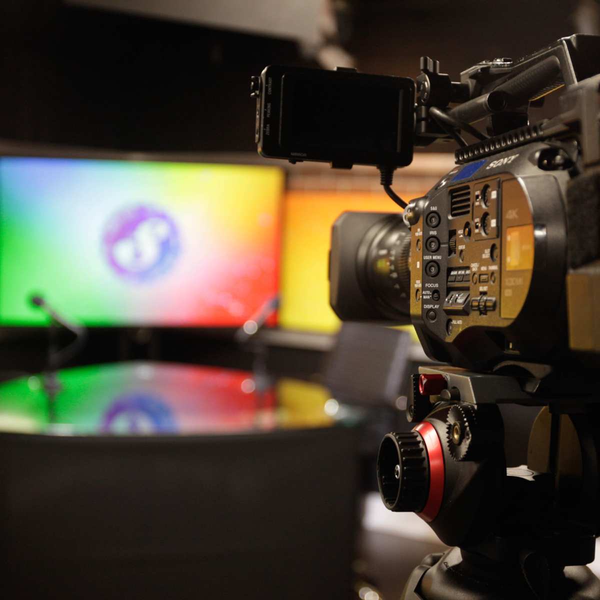 A video camera is positioned in front of a brightly lit television studio set with a circular table and a colorful backdrop.