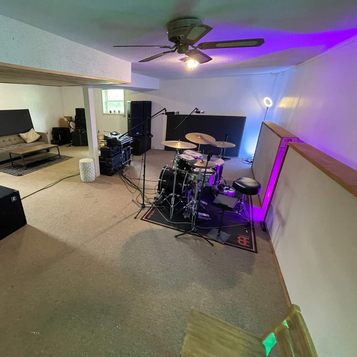 A basement space featuring a drum set and audio equipment, with a seating area visible in the background.