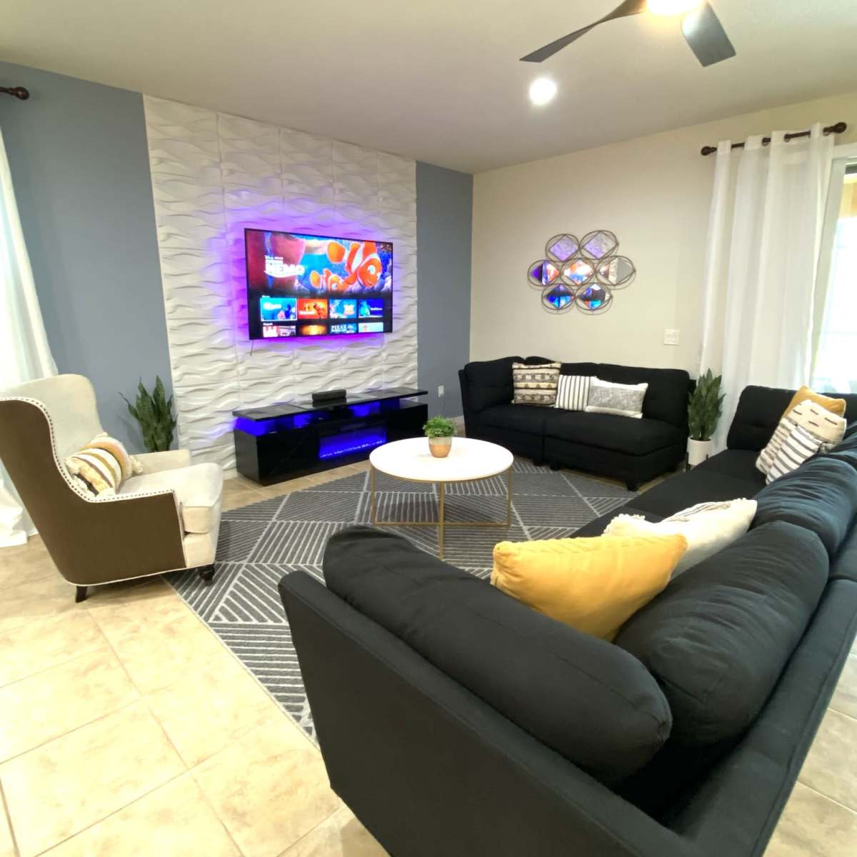 A living room features a sectional sofa, an armchair, a round coffee table, a television mounted on a textured wall, and decorative plants.