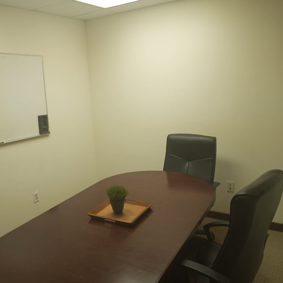 A small conference room features a wooden table with a plant centerpiece and two black chairs, alongside a whiteboard displaying handwritten notes.
