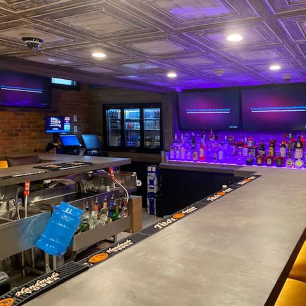 A bar area with a long countertop, illuminated bottles on shelves, and multiple screens displaying content in a dimly lit setting.
