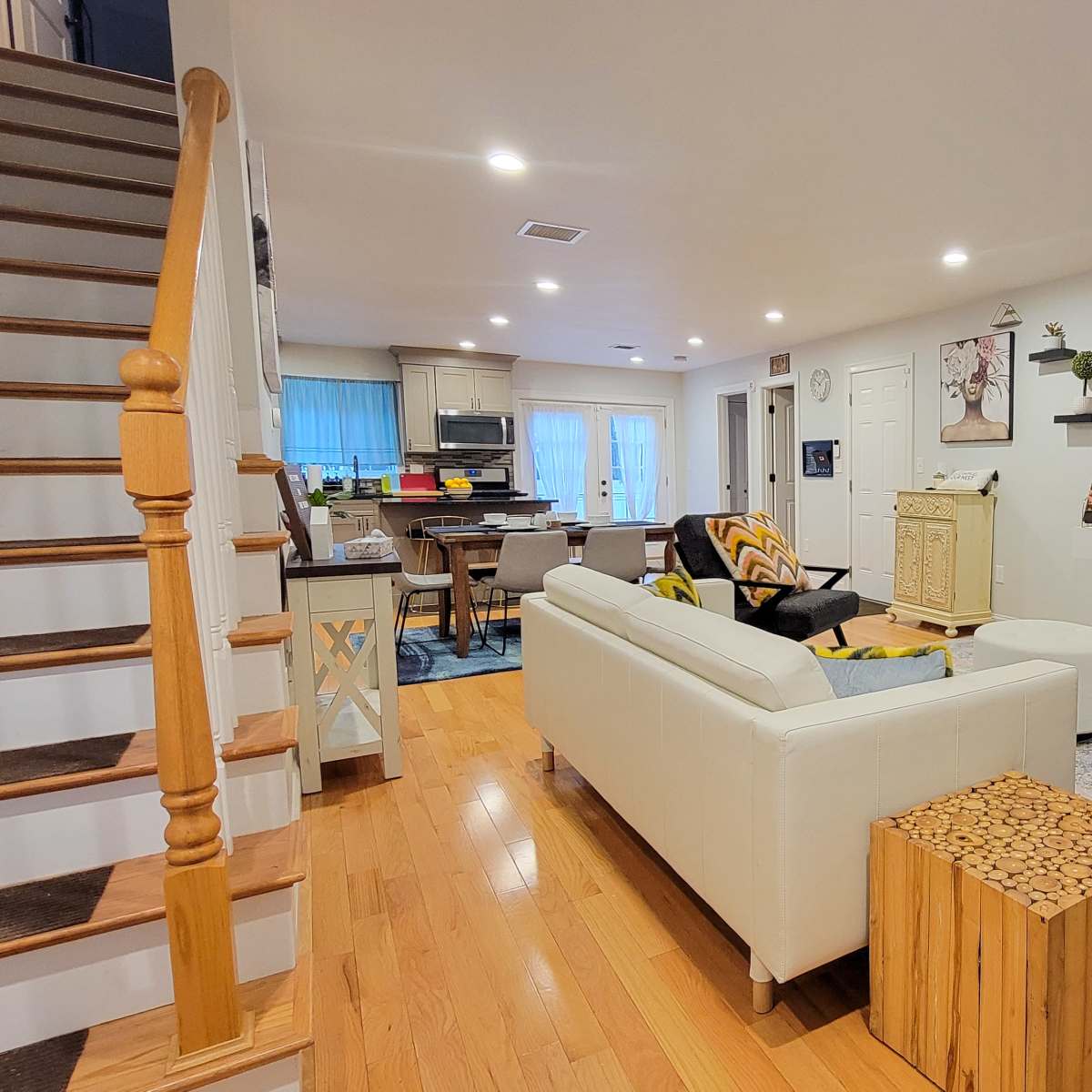 A modern living room with a staircase on the left, a sofa and armchair facing a wall-mounted television, and a dining area visible in the background.