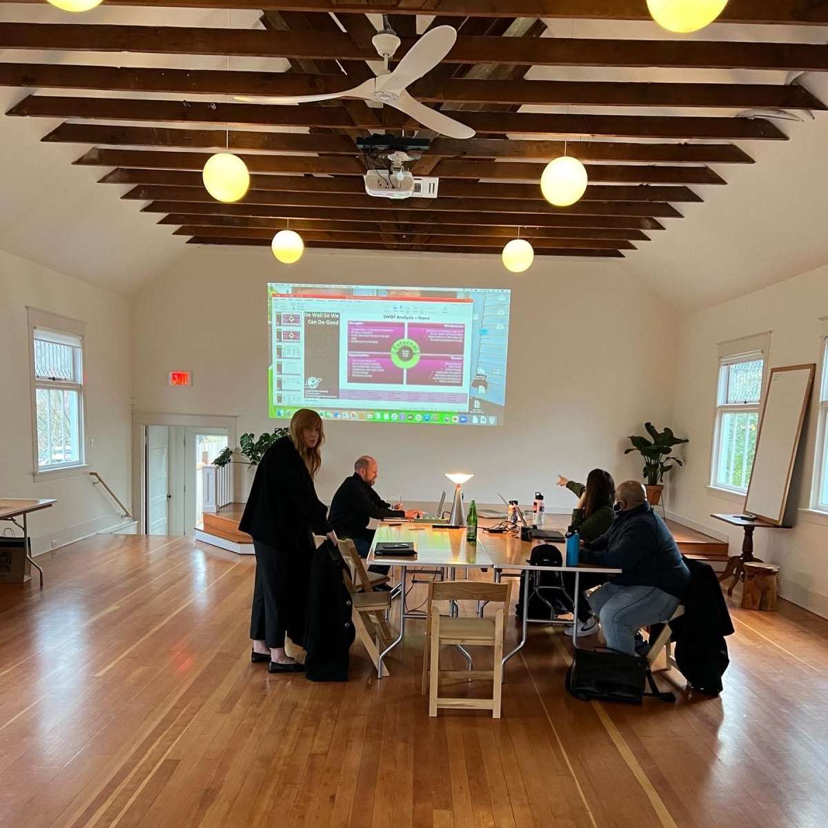 A group of people is gathered around tables in a spacious room with wooden floors and a large screen displaying a presentation.