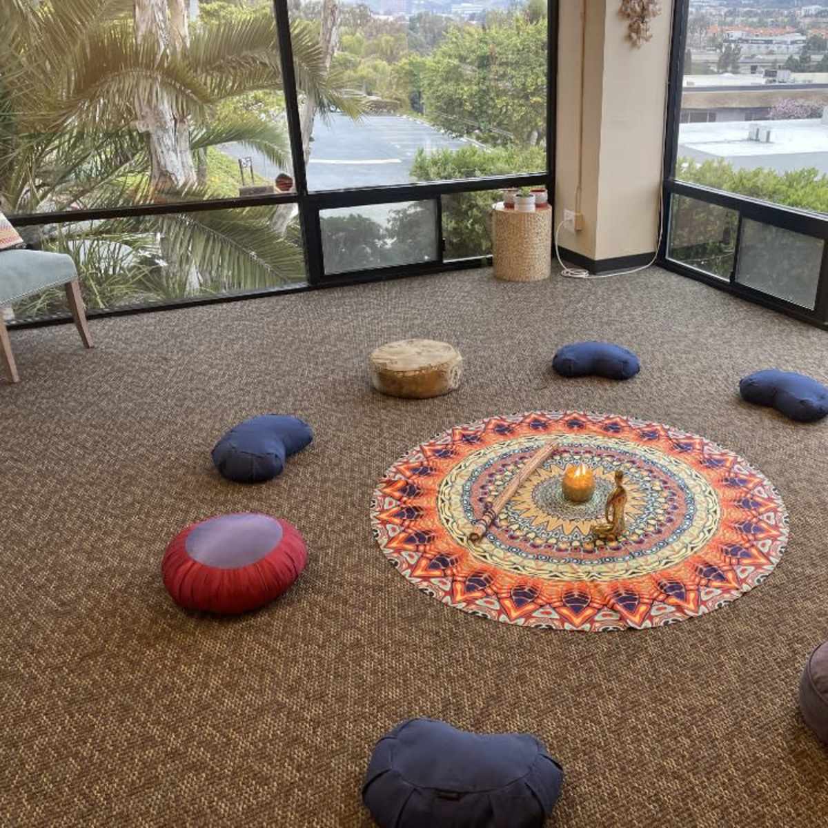 A circular arrangement of meditation cushions around a decorative mandala on the floor, set in a room with large windows overlooking a landscape.