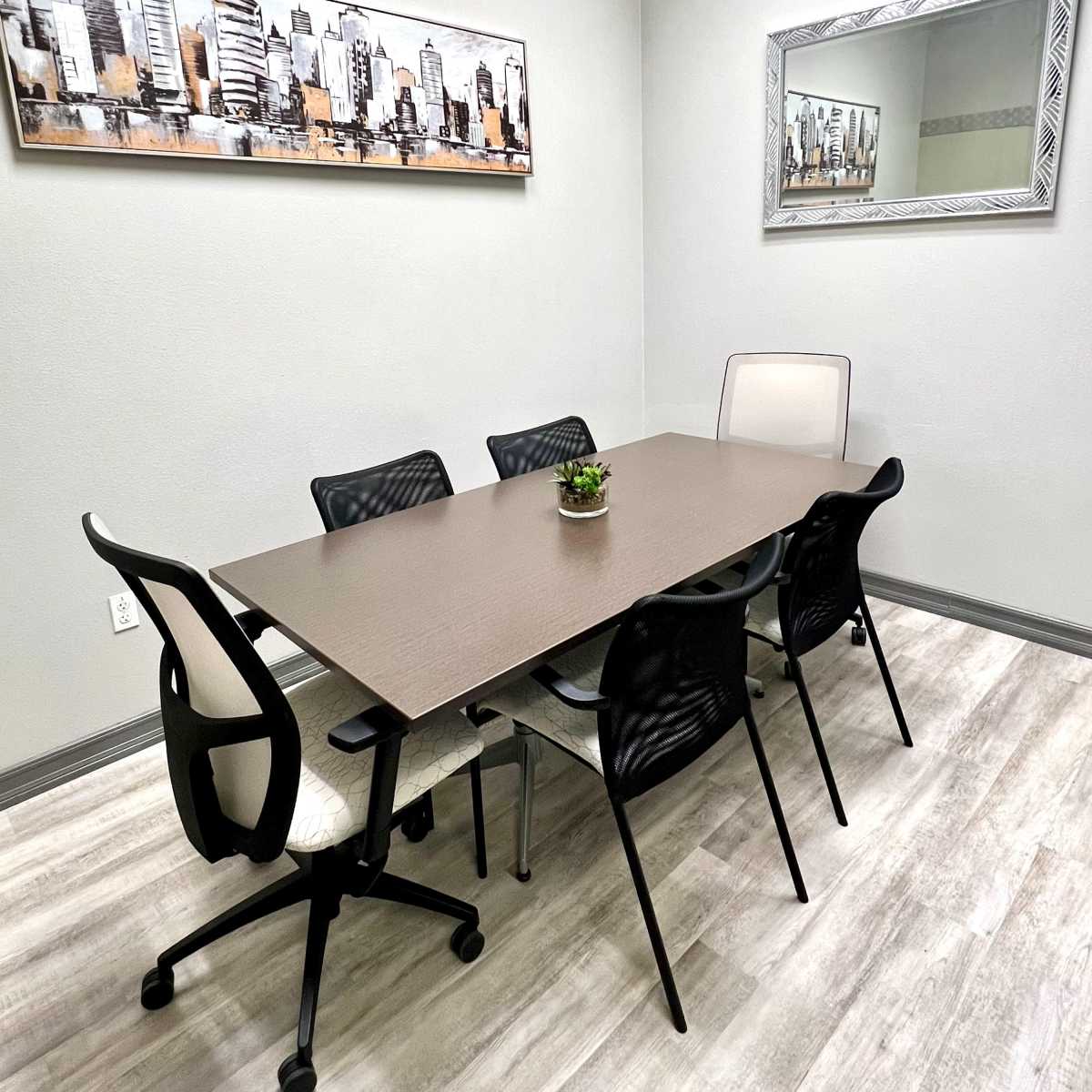 A small conference room with a rectangular table surrounded by six chairs, a wall-mounted cityscape art piece, and a mirror.