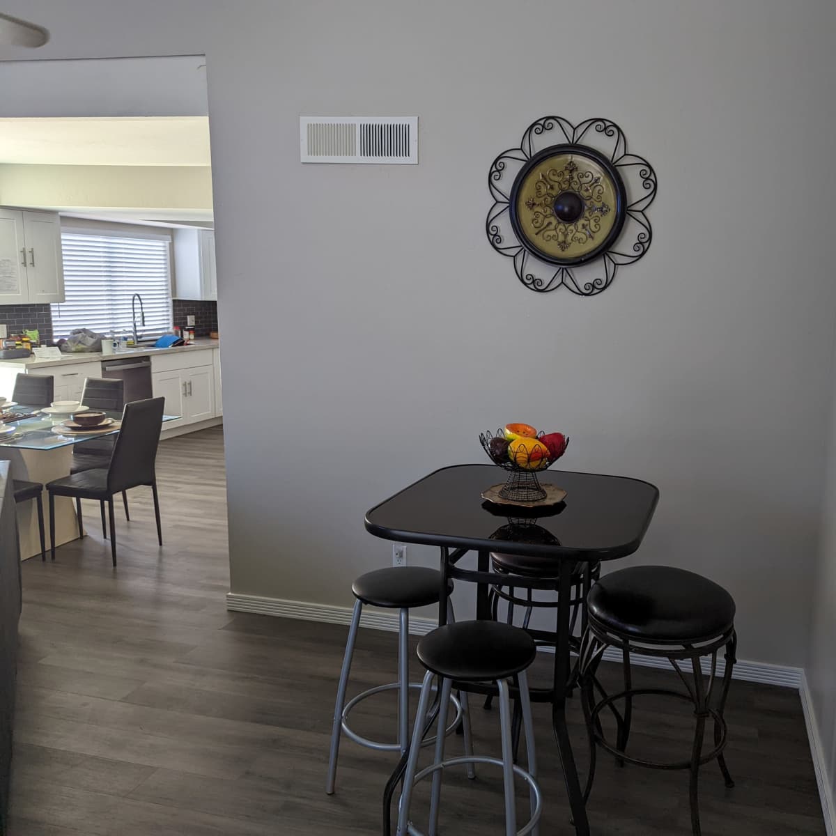 A small dining area features a round black table with four stools and a fruit bowl, situated against a light-colored wall decorated with a circular metal wall clock.