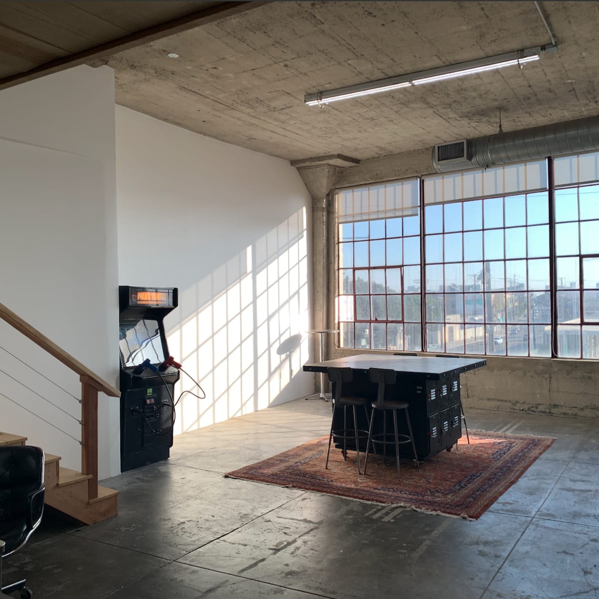 A spacious, industrial-style room with large windows, a staircase, a game machine, and a central table surrounded by stools on a patterned rug.