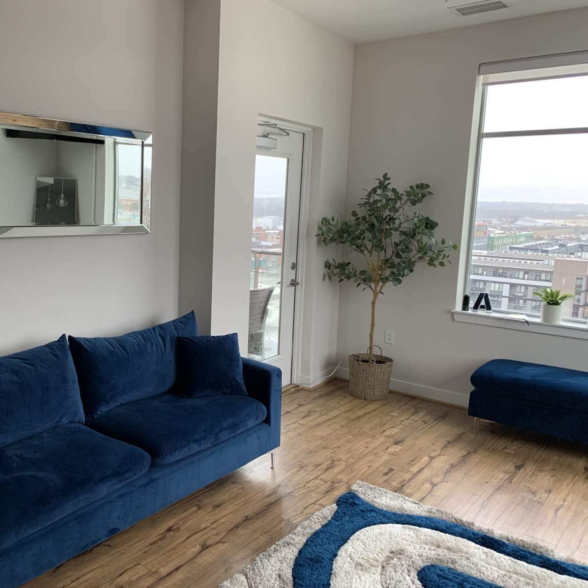 A blue sofa sits against a light wall beside a large mirror, with a potted plant and a window displaying a city view.