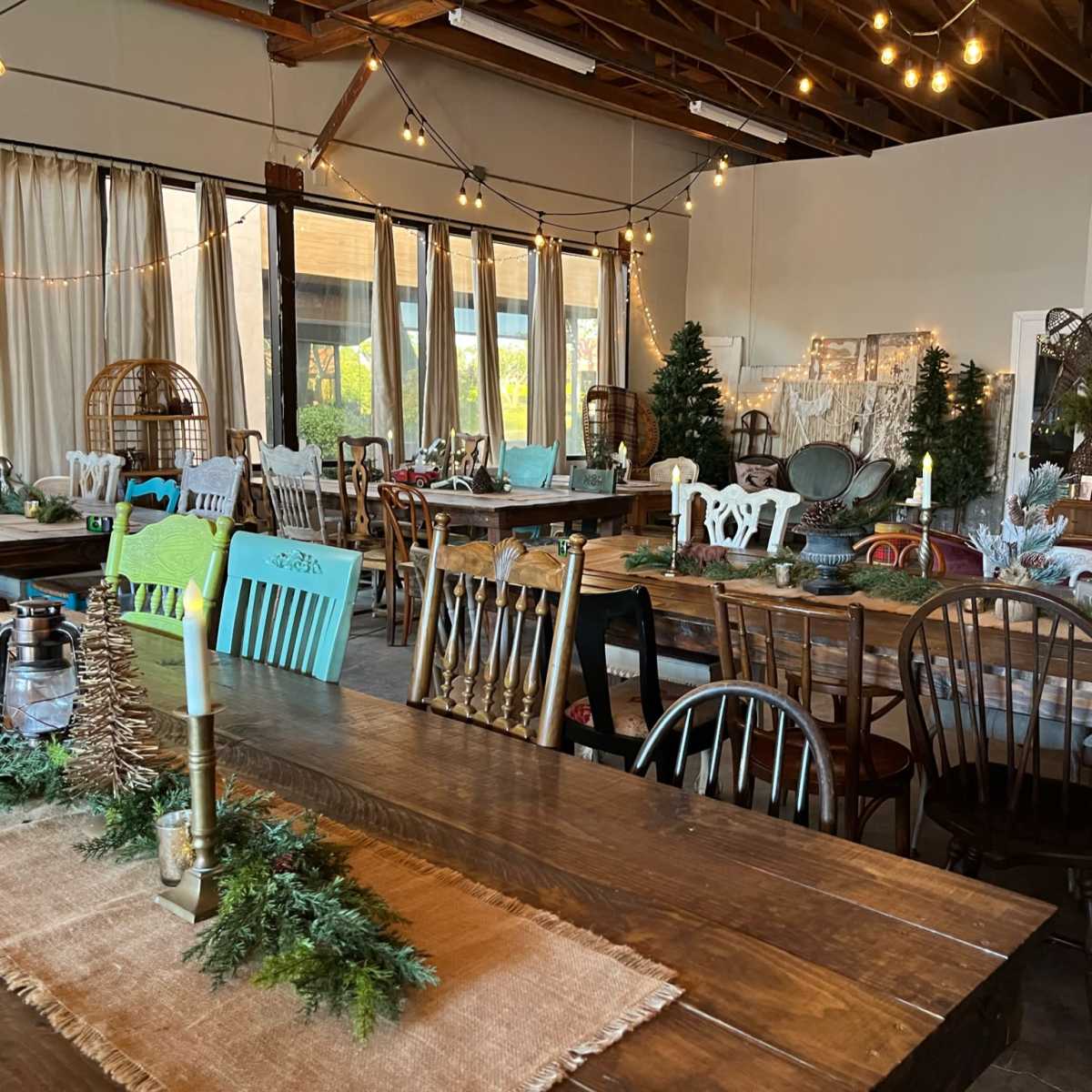 A spacious, decorated event room filled with various wooden tables and chairs, adorned with greenery and soft lighting.