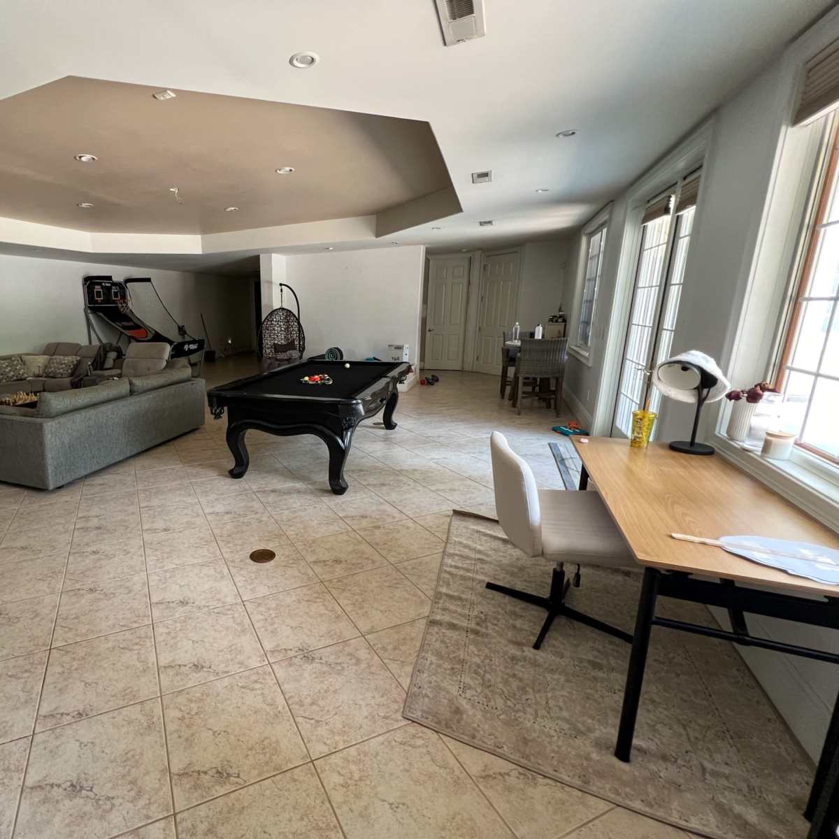 A spacious room featuring a pool table, a gray sofa, a dining table, and large windows with natural light.