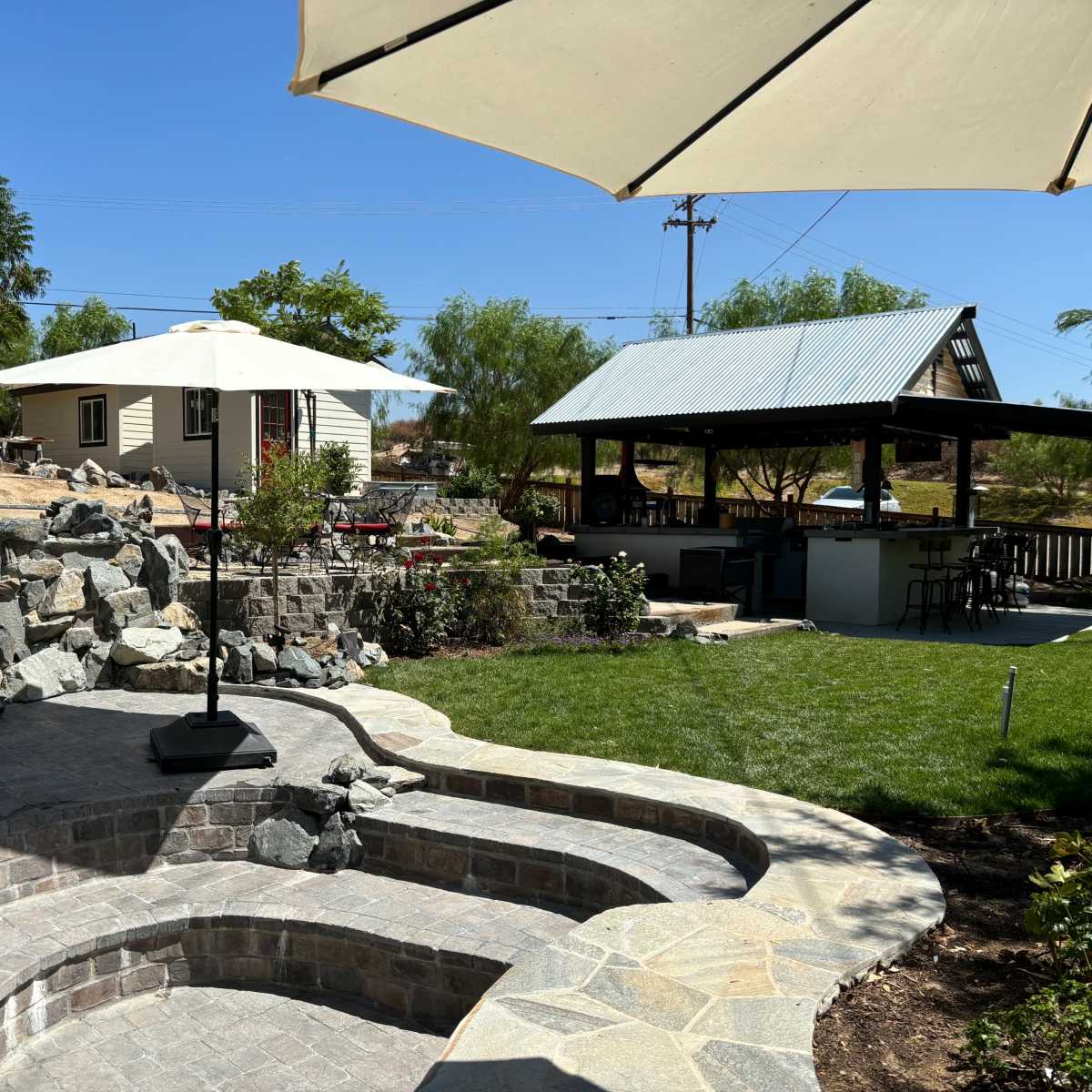 A patio area with stone steps, a red chair, and an umbrella, surrounded by a grassy lawn, a rock garden, and a small building in the background.
