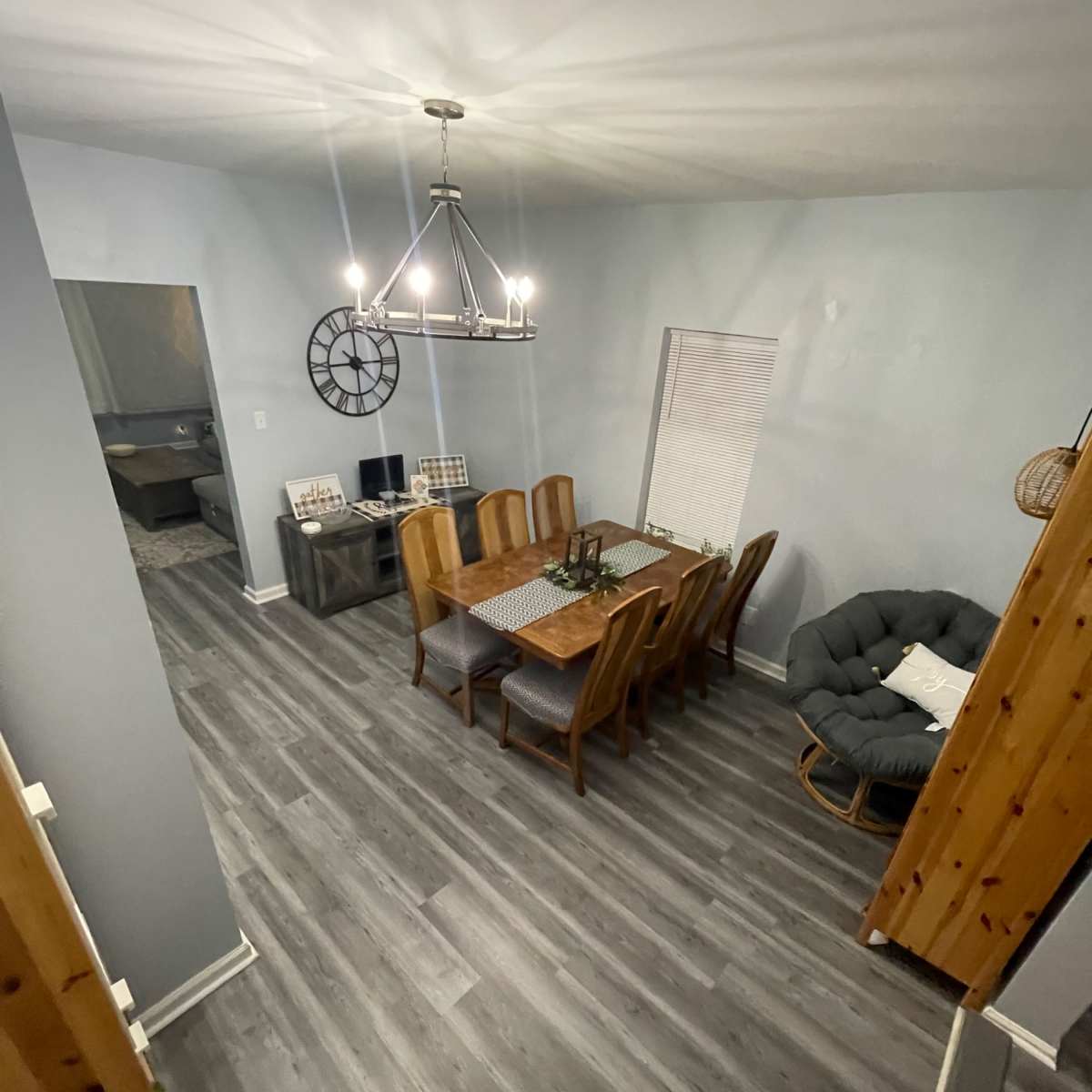 A dining area with a long wooden table surrounded by chairs, a chandelier above, and gray walls and flooring.
