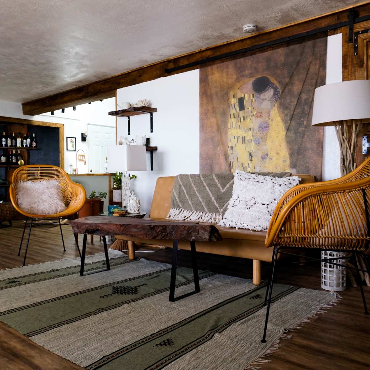 A cozy living area featuring a brown leather sofa, wicker chairs, a wooden coffee table, and a patterned rug, with a large artwork on the wall and a bar area in the background.