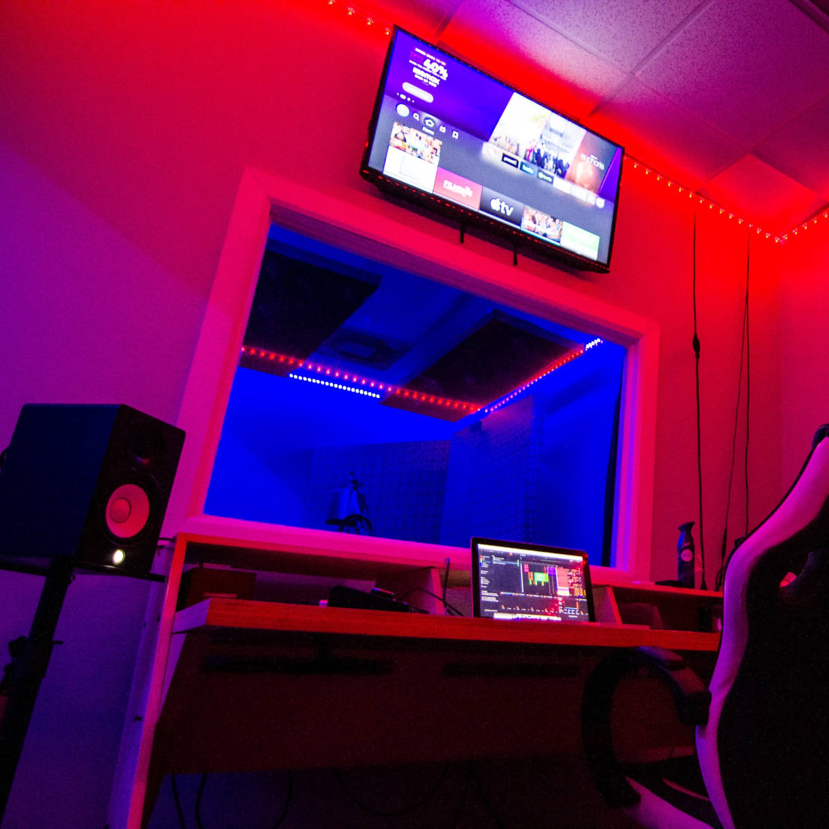 A recording studio features a desk with a computer and an ergonomic chair, illuminated by red and blue LED lighting, with soundproof panels on the walls and a TV mounted above.