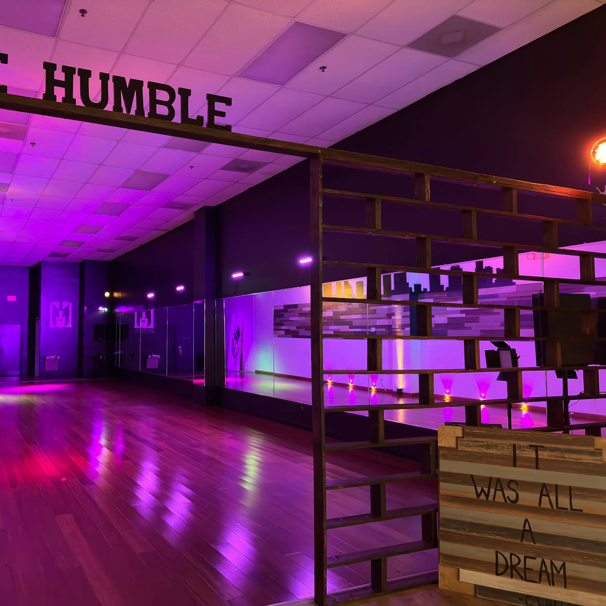 A dance studio with colorful lighting, decorated with the words "BE HUMBLE" at the entrance and a sign that reads "WAS ALL A DREAM" on a wooden structure.