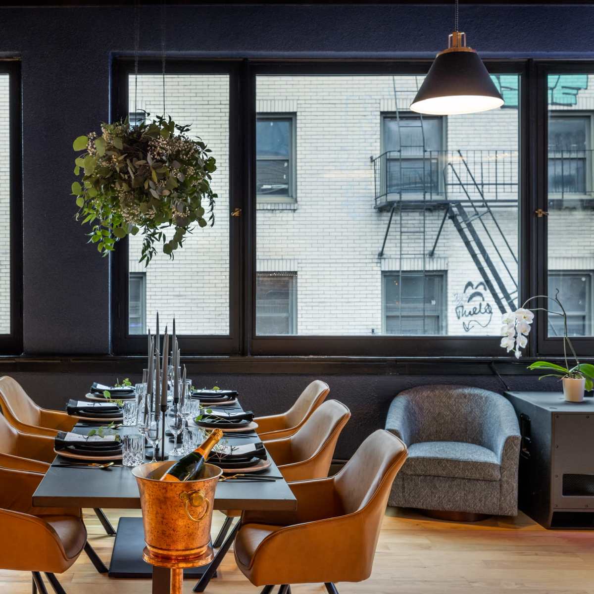 A dining table is set with plates and glasses, surrounded by modern chairs, in a well-lit room with large windows overlooking a brick building.