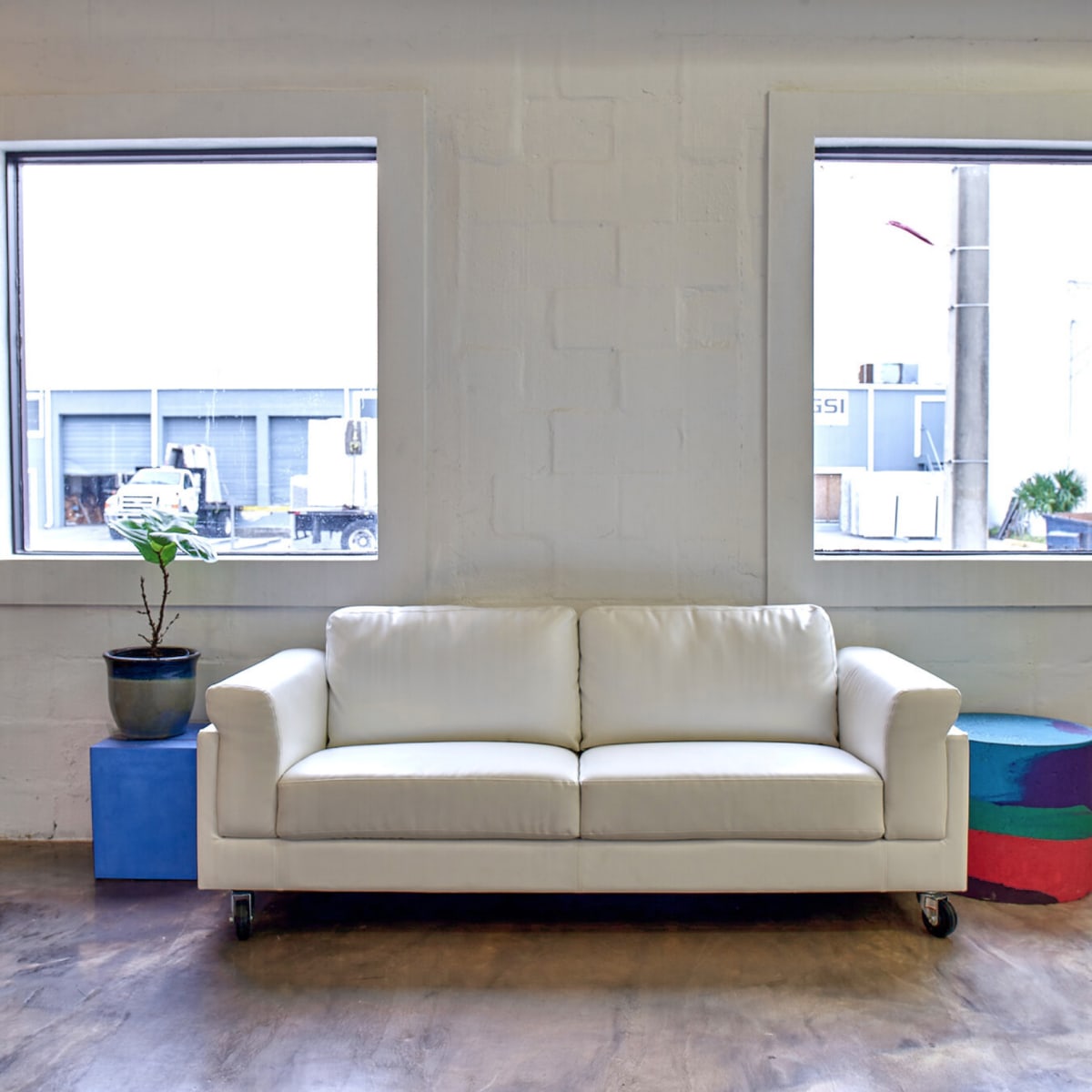 A white sofa sits in a room with two large windows, alongside a small potted plant and colorful side tables.