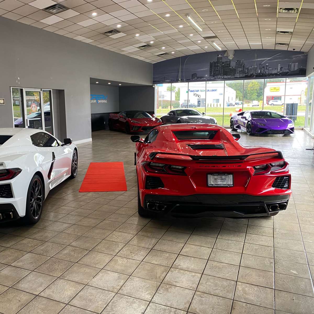 A car dealership interior featuring a white Corvette and a red Corvette in the foreground, with additional luxury cars visible in the background.