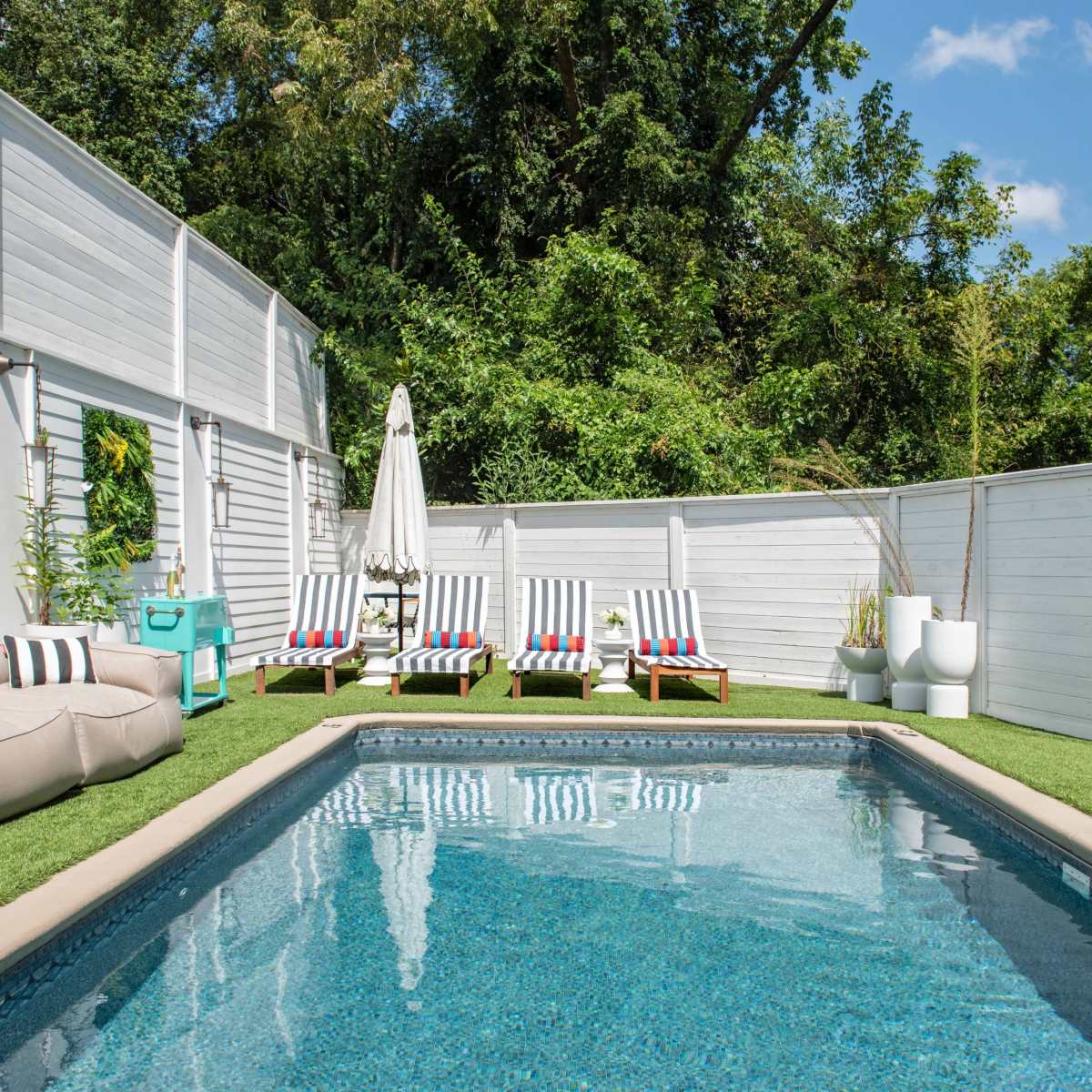 A swimming pool surrounded by lounge chairs and a neatly landscaped outdoor area with greenery and a white privacy fence.