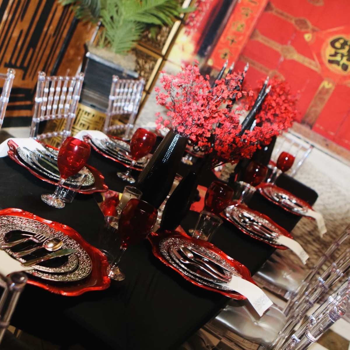 A dining table is set with ornate dishware and red floral arrangements against a backdrop of a red door with traditional motifs.