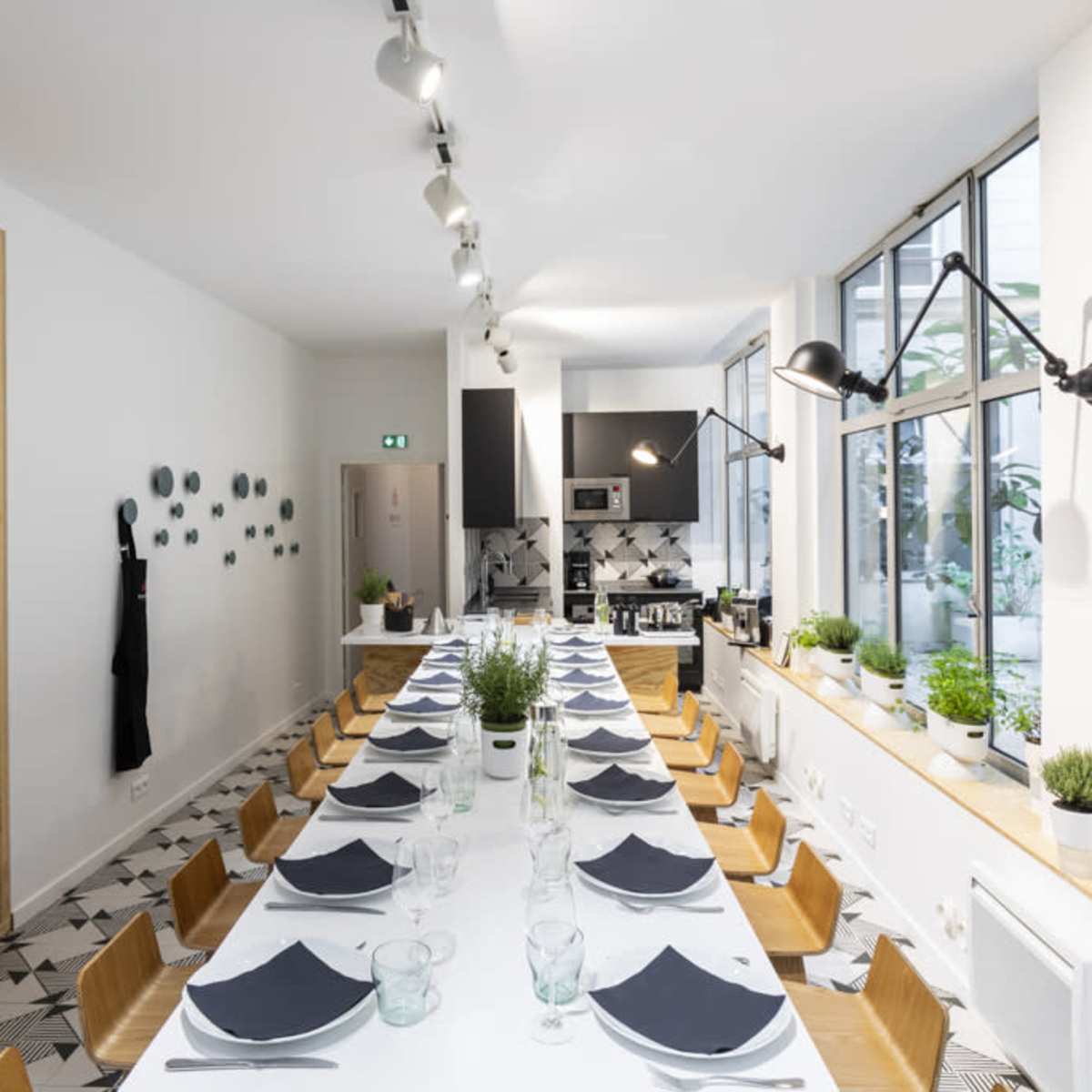 A long dining table is set with dark napkins and glassware in a bright, modern kitchen featuring large windows and minimalist decor.