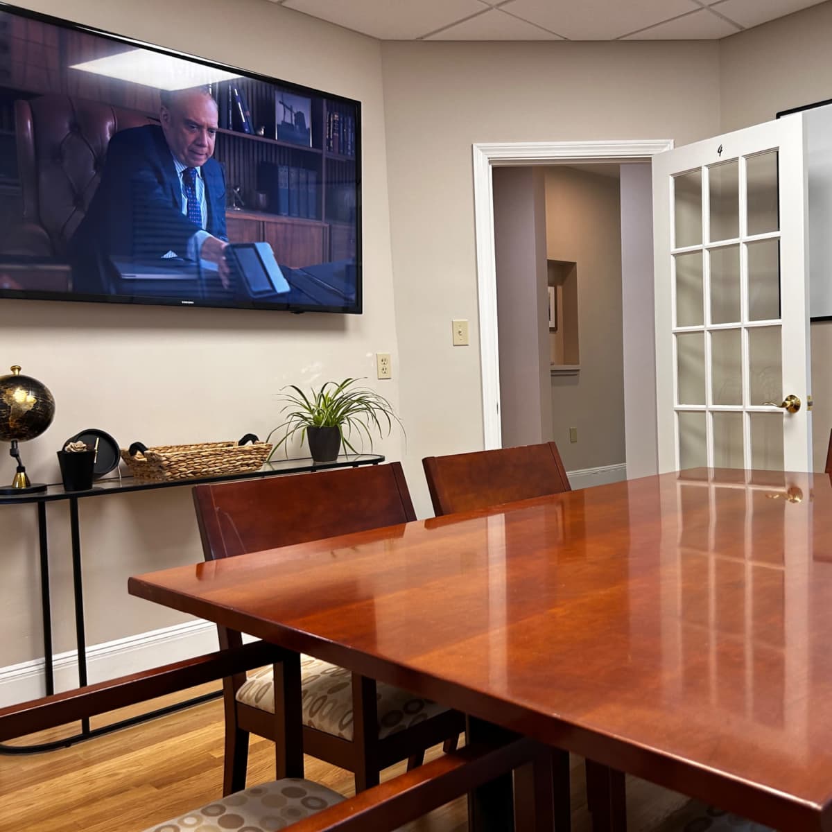 A conference room with a wooden table, chairs, a screen displaying a man, and a whiteboard on the wall.