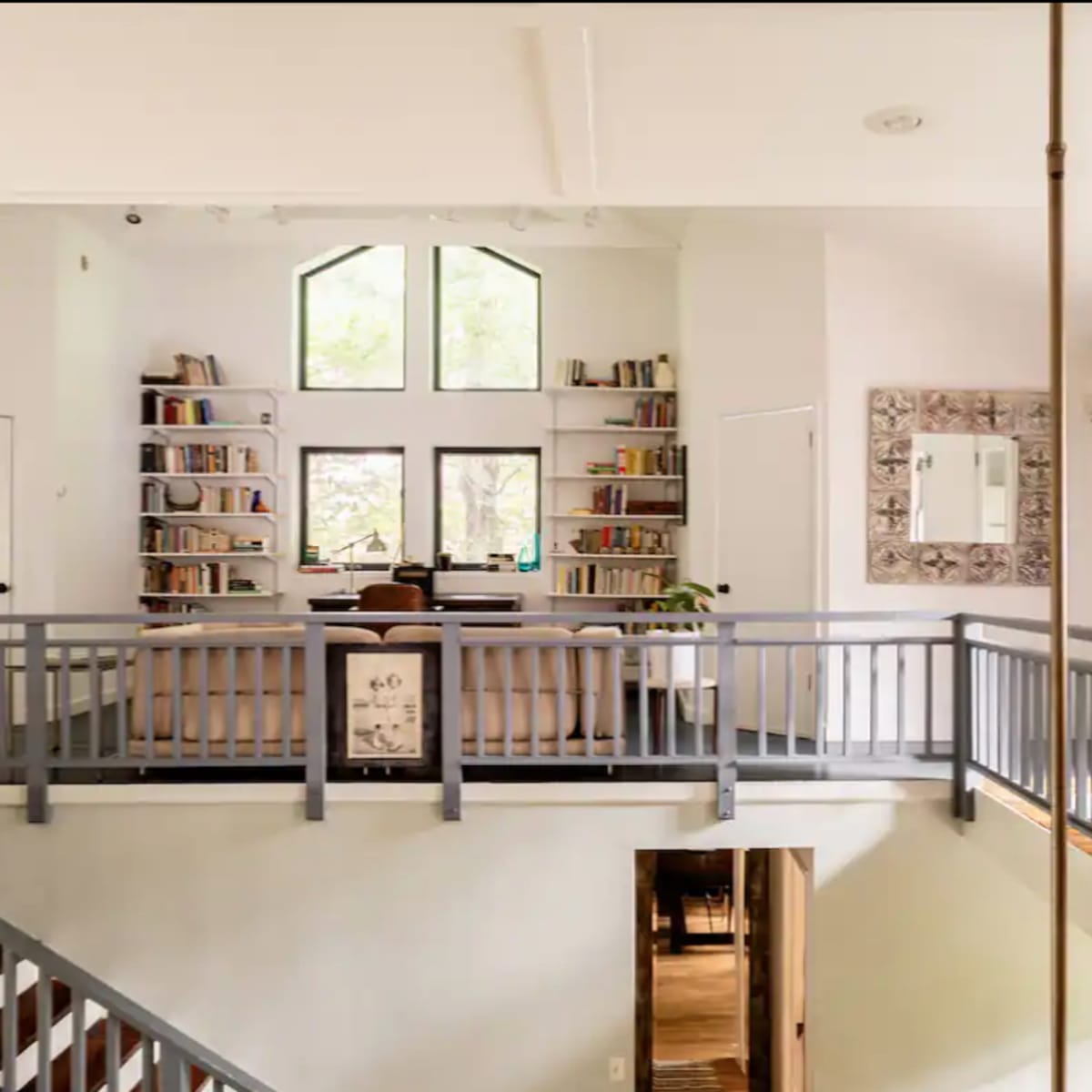 A two-level living space with a bookshelf-filled wall, a couch, and large windows allowing natural light to enter.