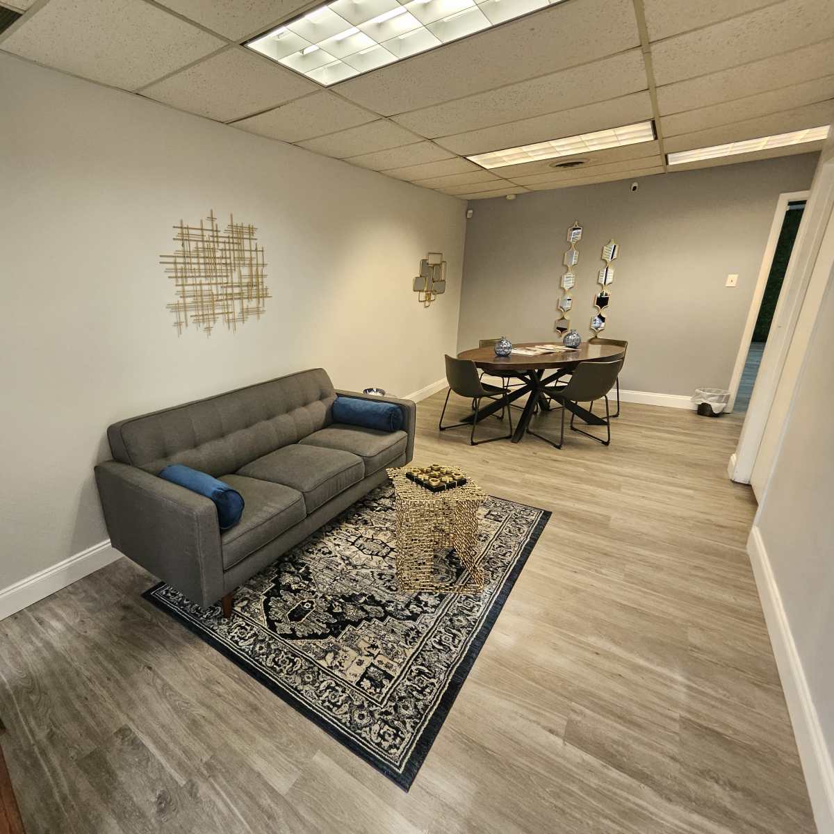 A small office space featuring a gray sofa, a patterned area rug, a wooden table with chairs, and decorative wall art.