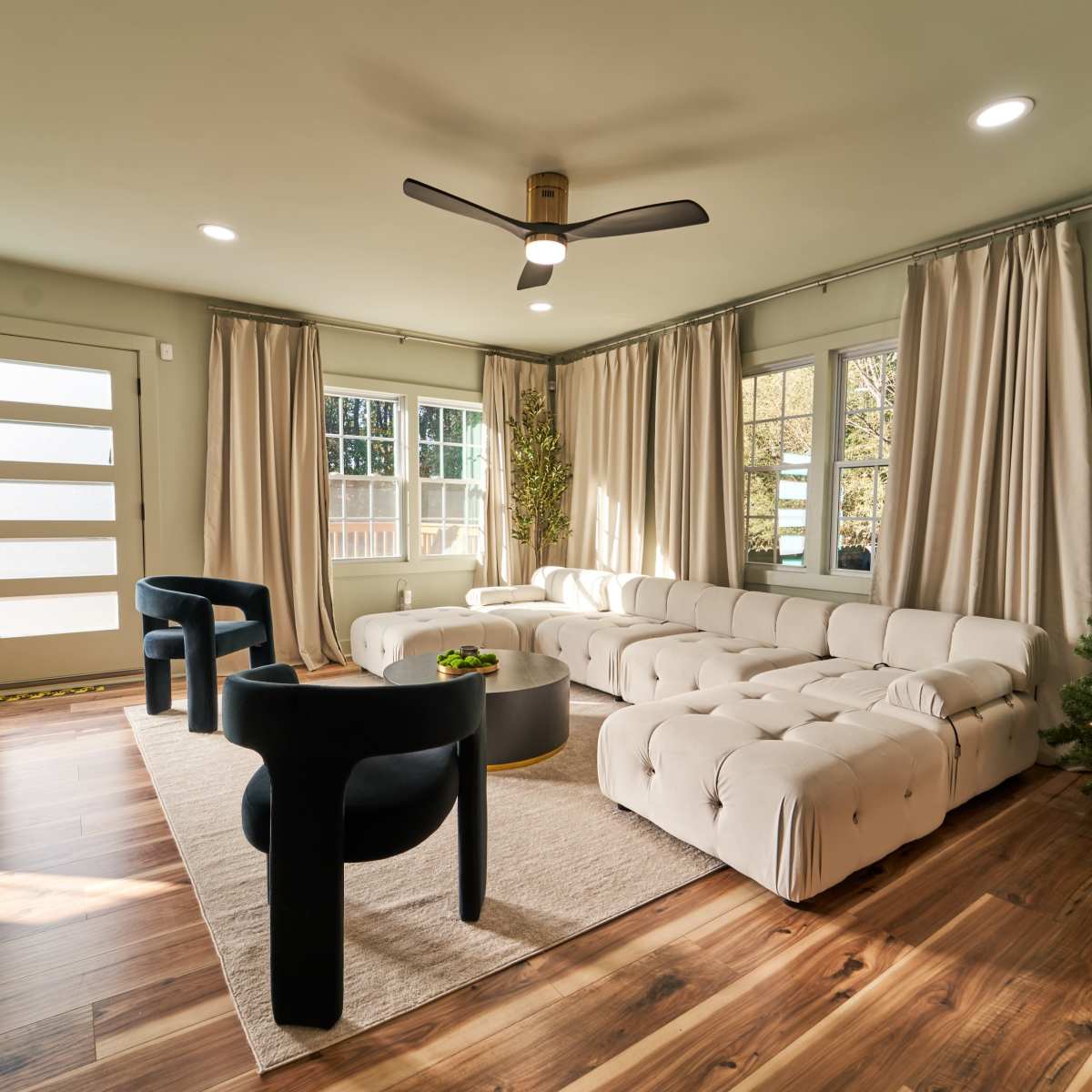 A modern living room features a beige tufted sectional sofa, two black accent chairs, a round coffee table, and a Christmas tree, with large windows and light-colored curtains.