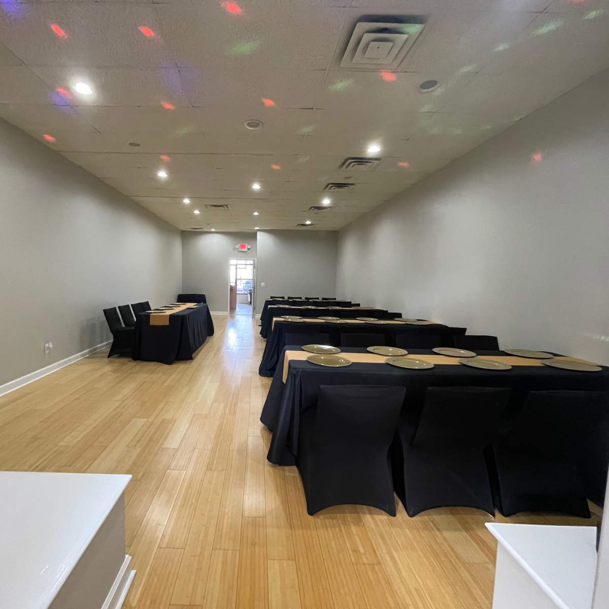 A long, narrow event space with black tablecloths covering several tables, illuminated by multicolored lights on the walls and ceiling.