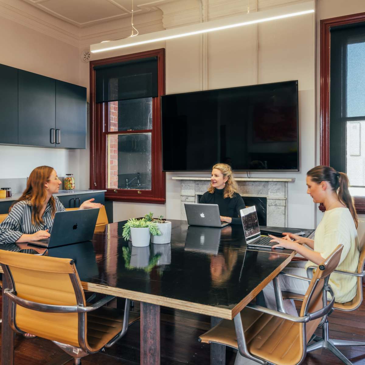Three women are seated at a conference table in a modern office with laptops, discussing while a large screen displays a view of the room.