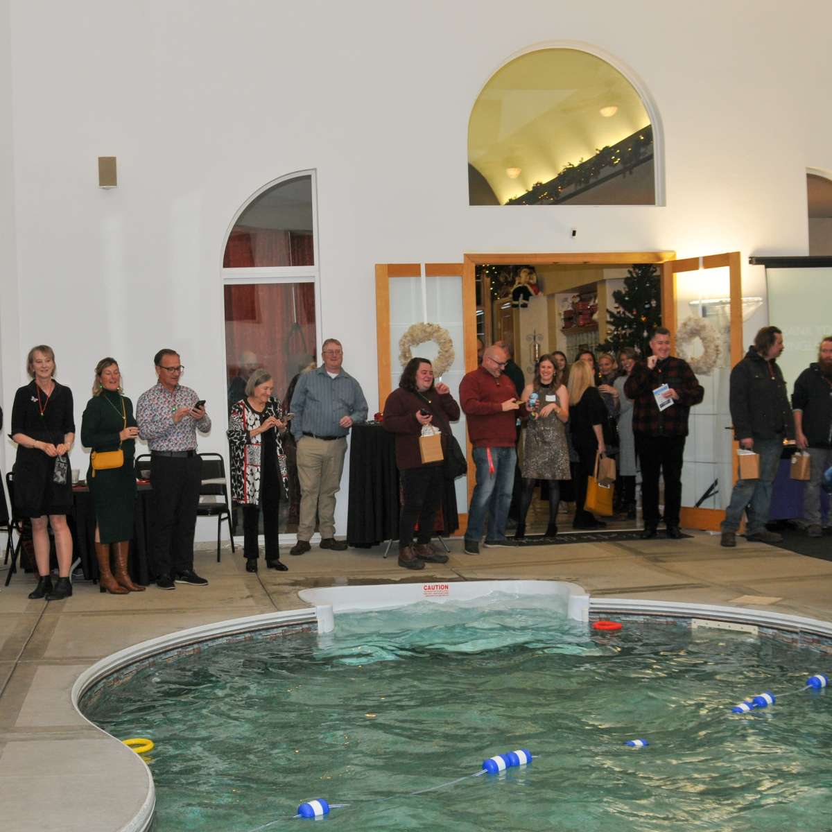 A group of people gathers around a swimming pool in a spacious indoor venue, with some standing and others seated in chairs along the walls.