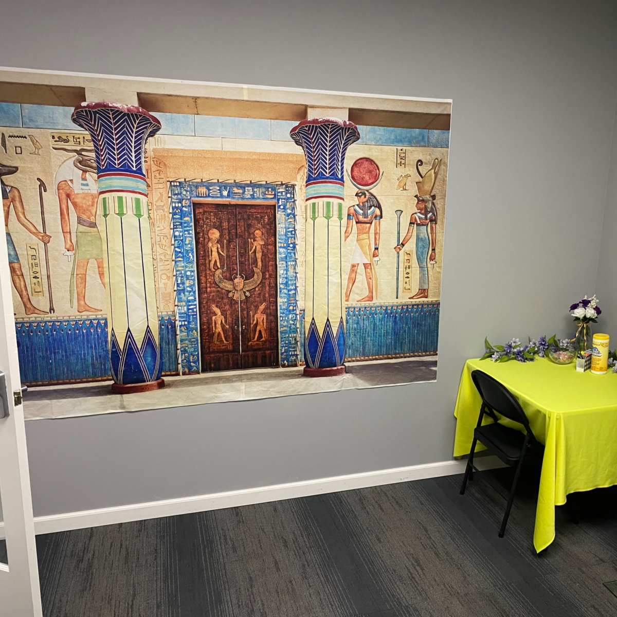 A room with a large mural of an ancient Egyptian entrance on one wall, a table covered with a bright yellow cloth, and a mannequin dressed in a patterned gown on the right.