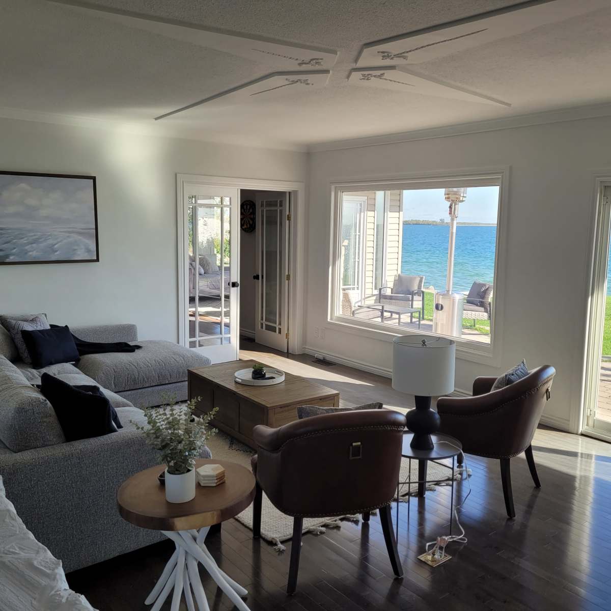 A modern living room features a gray sofa, two brown armchairs, and large windows offering a view of the water.