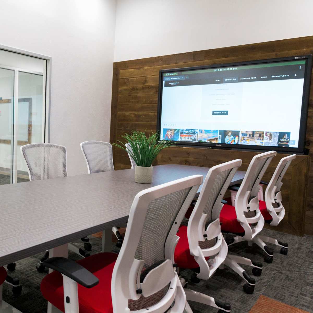 A conference room features a large table surrounded by white and red chairs, with a screen displaying a web page on the wall.