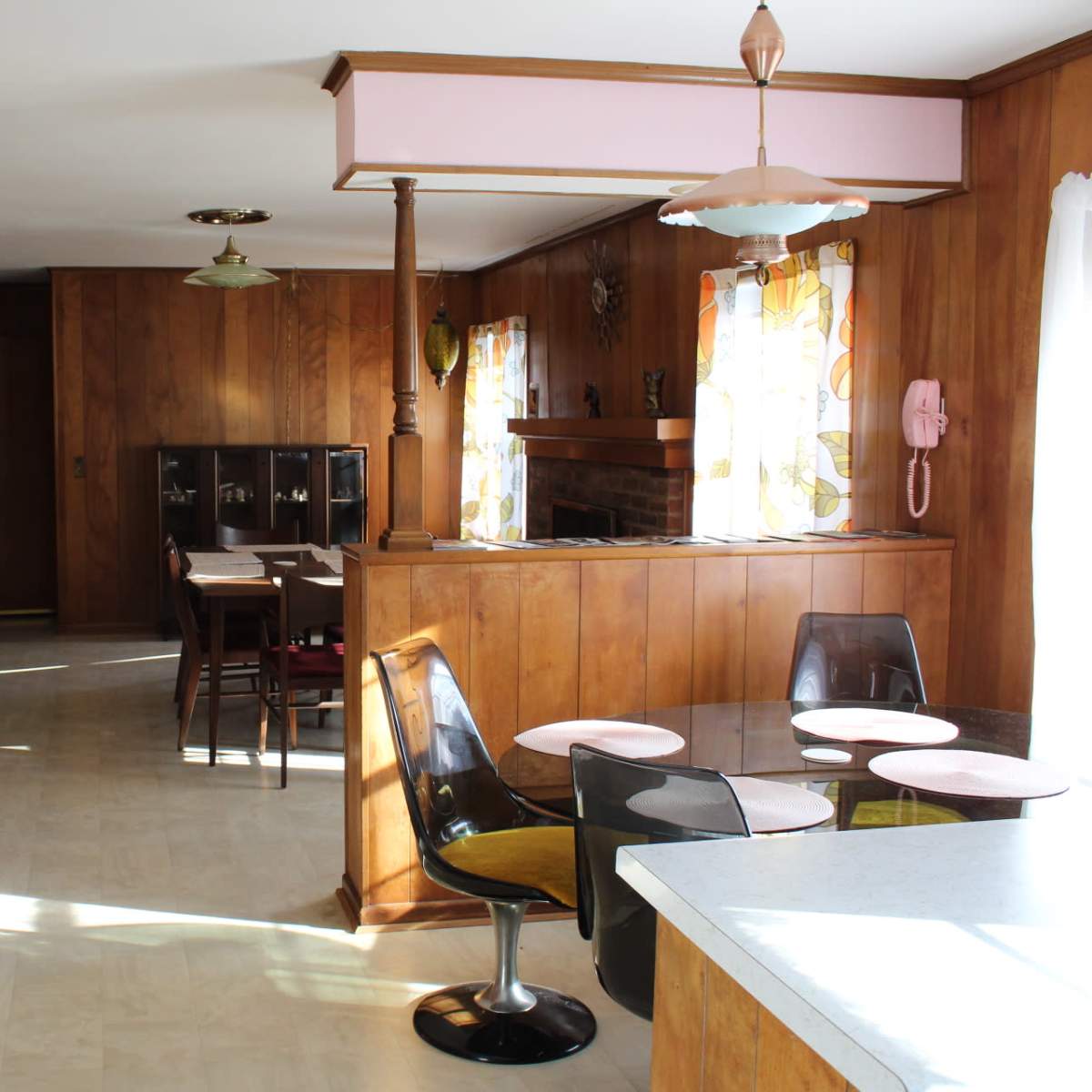 A spacious kitchen with wooden paneling, featuring a round dining table with chairs and an adjacent dining area with a large window adorned with colorful curtains.