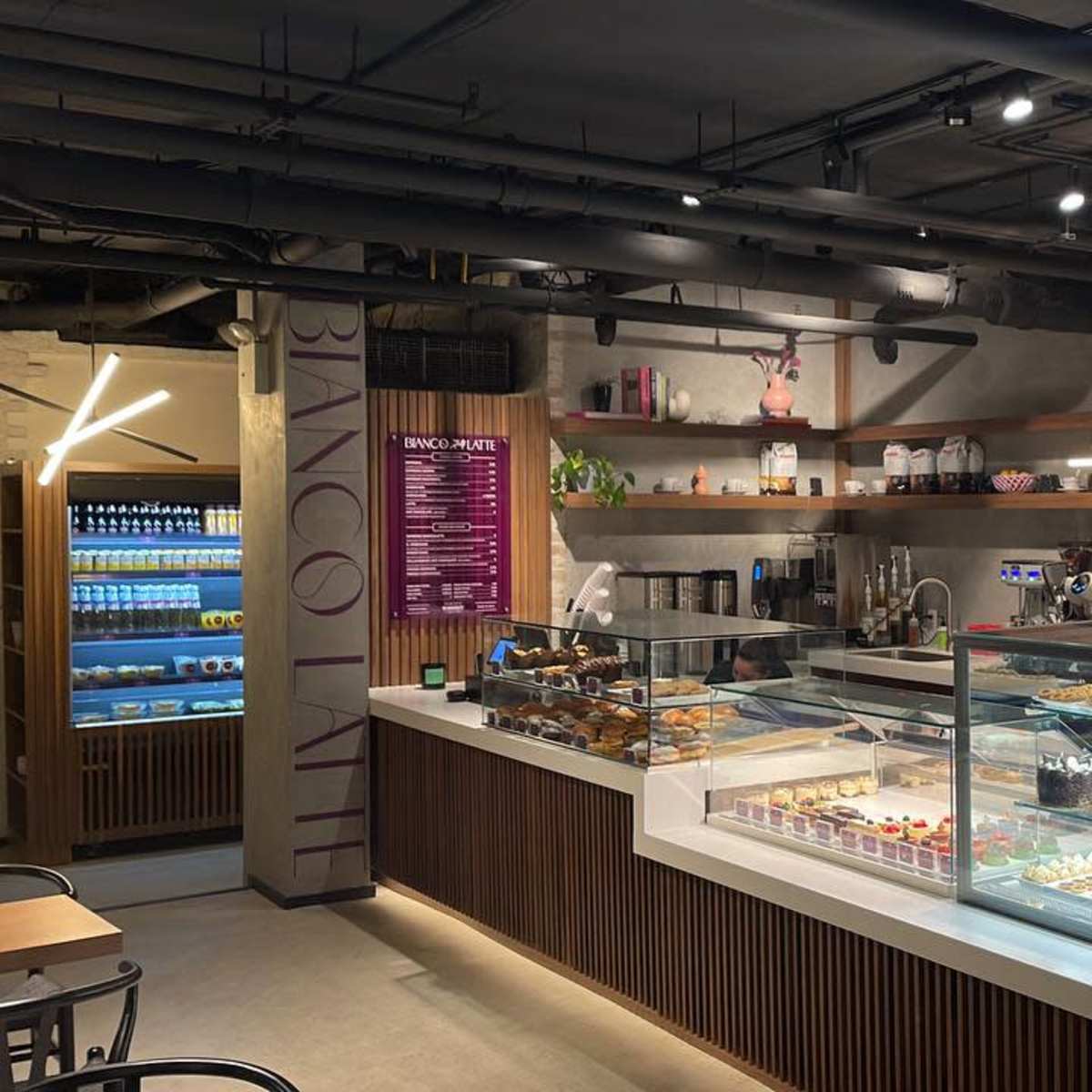 A modern café interior features a display case with an assortment of pastries and desserts, along with shelves holding various coffee-making equipment and drinks.