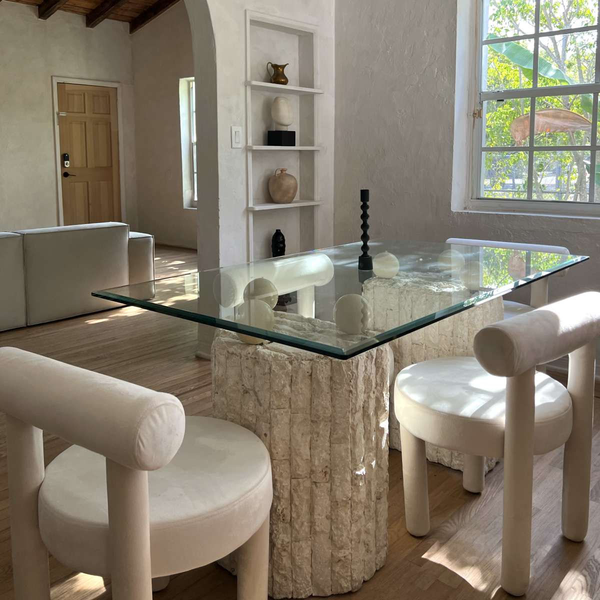 A dining area with a glass table supported by a textured stone base, surrounded by four tubular chairs, in a room with light-colored walls and wood accents.