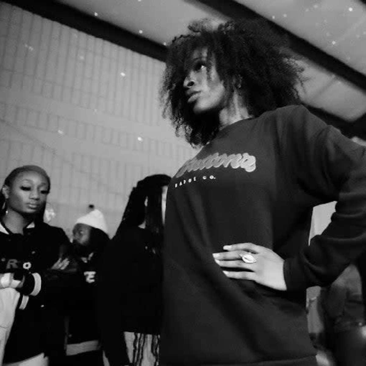 A model poses confidently in a black sweatshirt while spectators gather in the background.