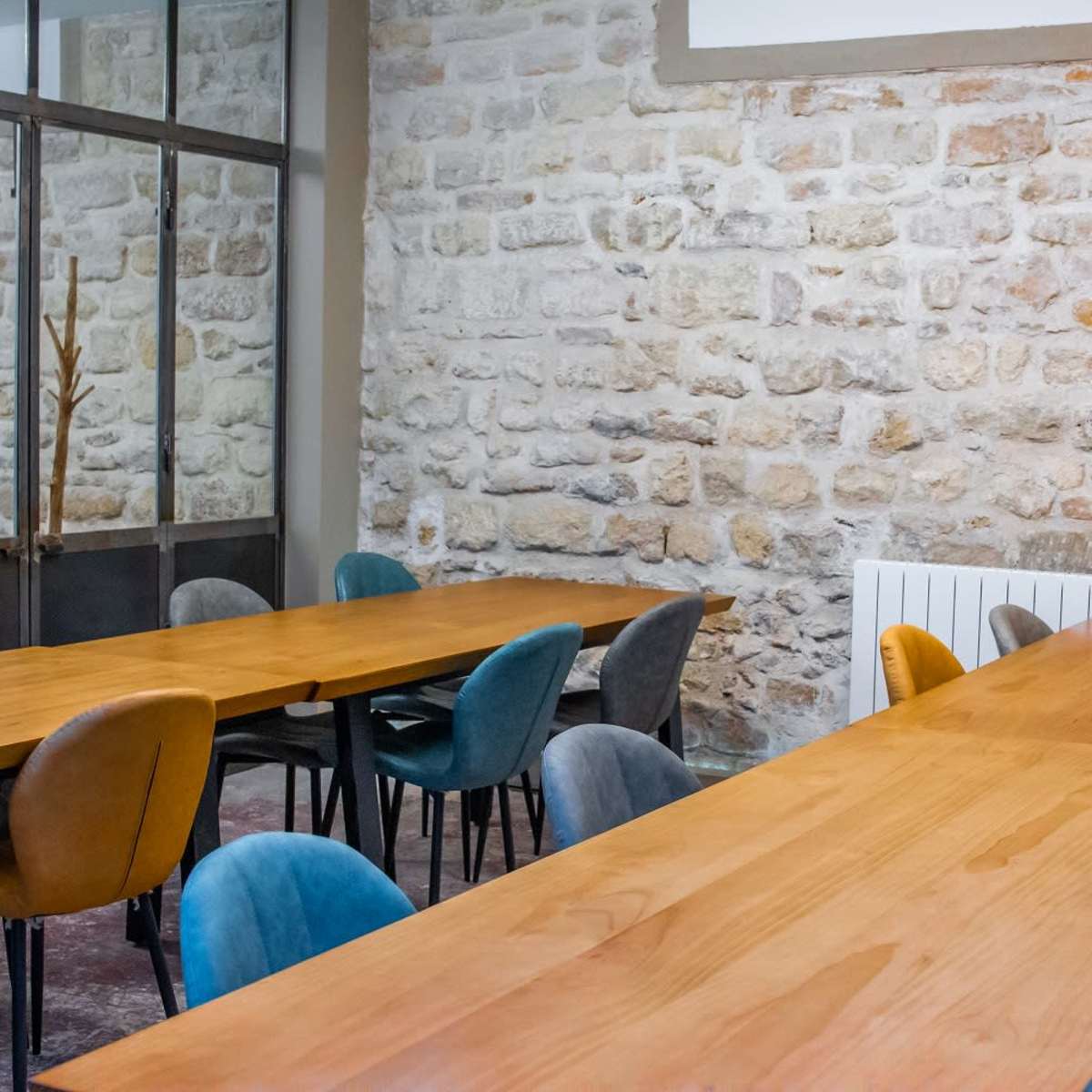 A meeting room features multiple long wooden tables surrounded by colorful chairs, with a stone wall in the background.