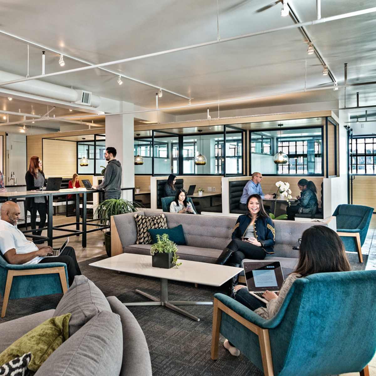 A modern office space with various groups of people engaging in different activities, including conversations, working on laptops, and standing at a bar-like counter.