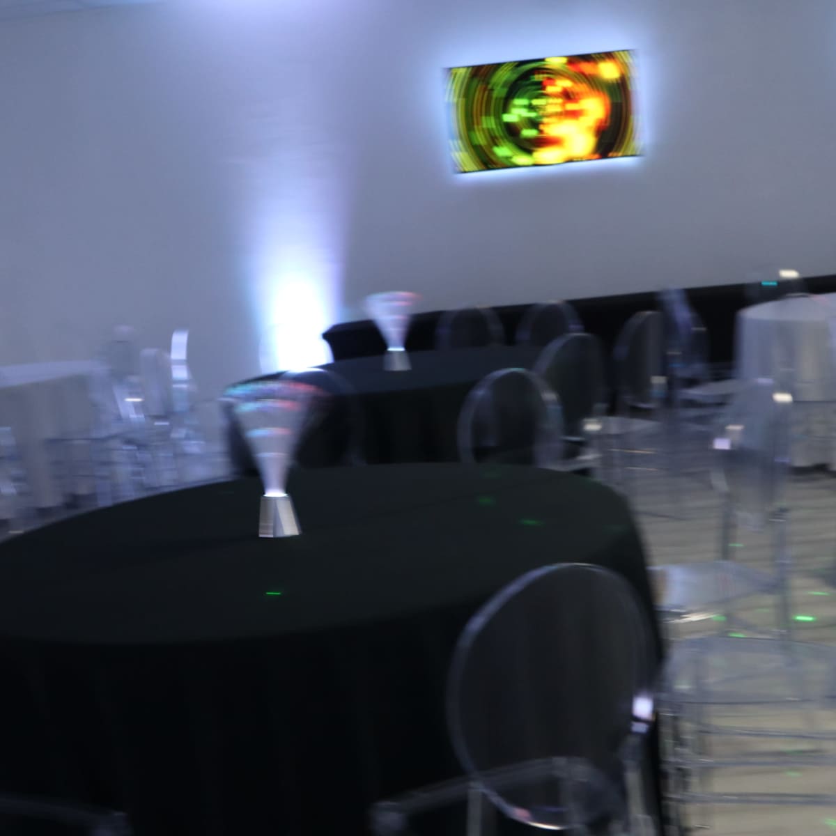 An event space with round tables covered in black linens and transparent chairs, illuminated by colored wall displays.