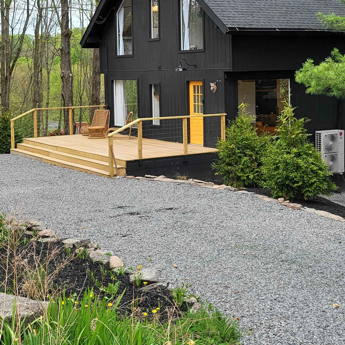 A black wooden house with a yellow door features a front deck and is surrounded by greenery and gravel pathways.
