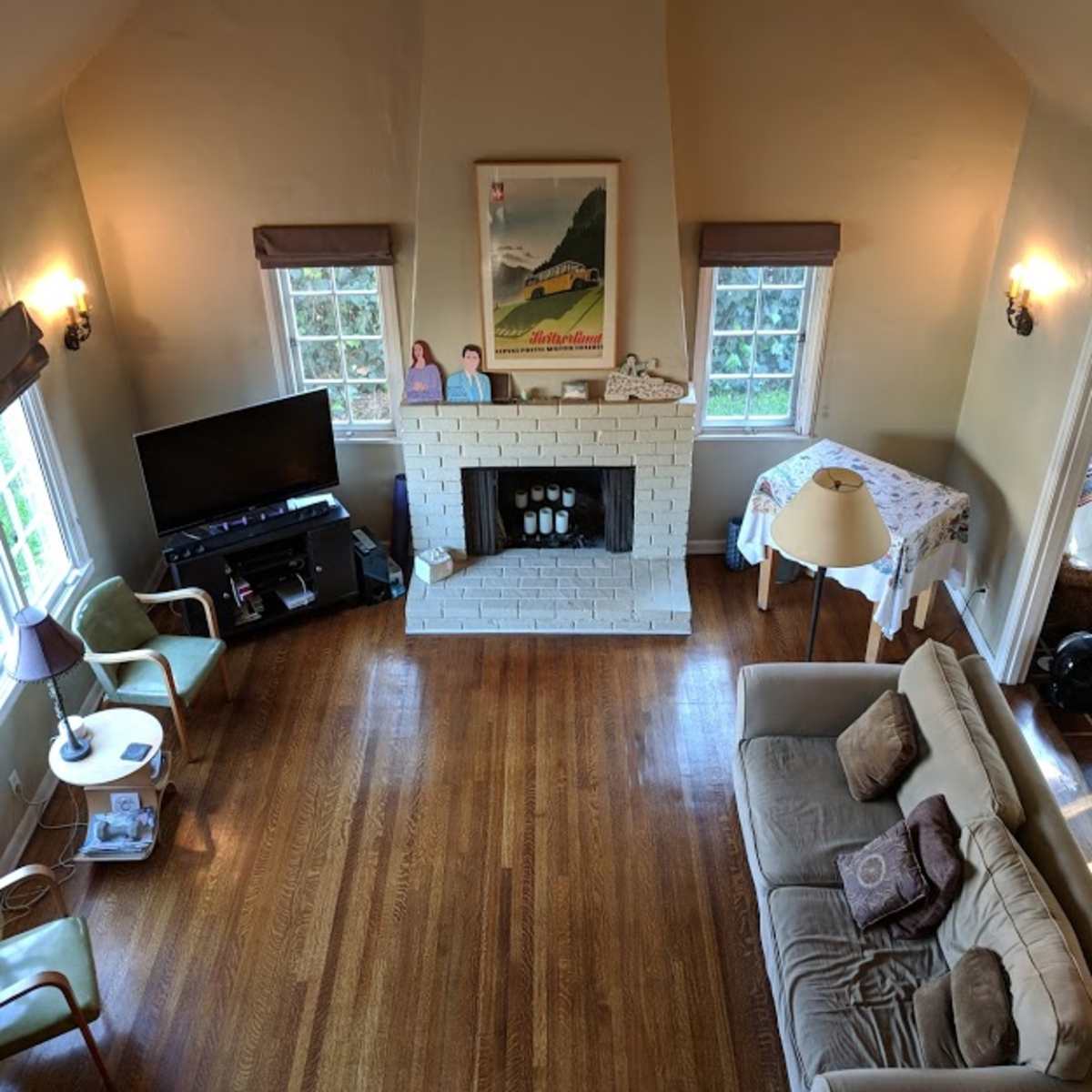 An interior of a living room with high ceilings, a white brick fireplace, two windows, a television, and a beige sofa.