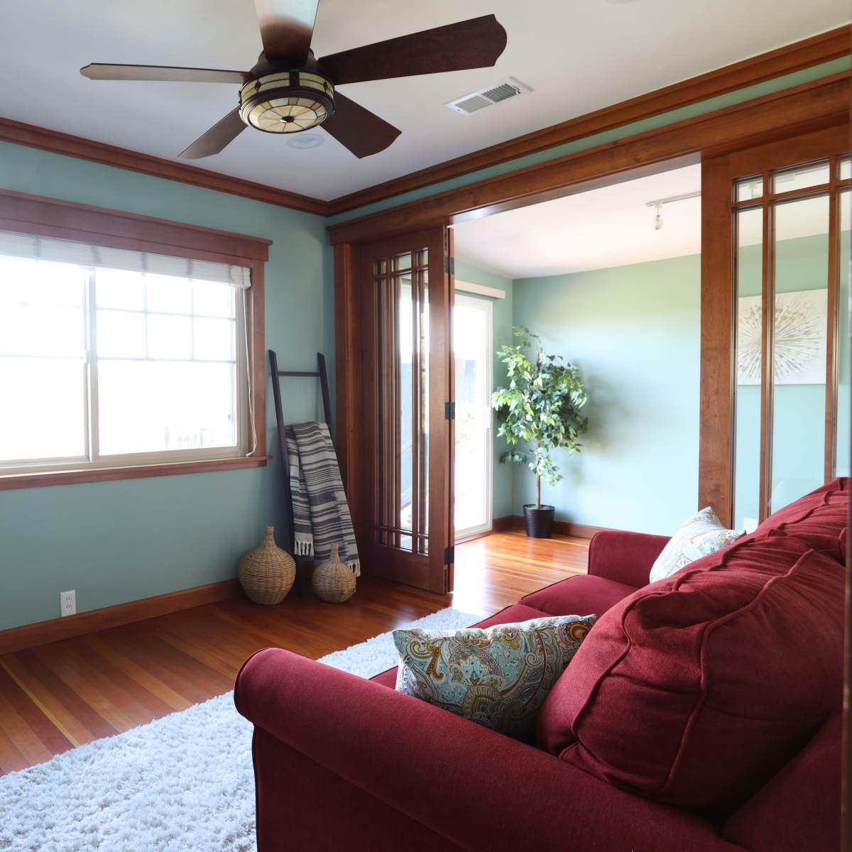 A cozy living room features a red couch, a wooden ceiling fan, and large windows with natural light.
