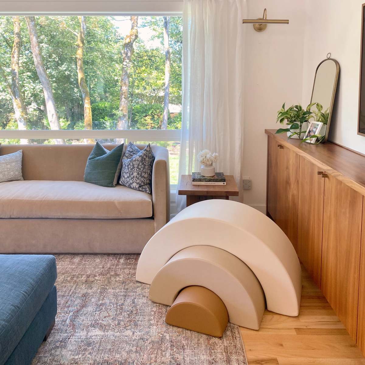 A cozy living room features a beige sofa, a wooden sideboard, and a stack of sculptural, curved decor in neutral tones near a large window.